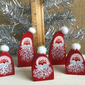 Pode incluir: Cinco ornamentos de madeira de Papai Noel vermelhos com pompons brancos no topo. Cada Papai Noel tem uma barba branca e um rosto branco com olhos pretos e um nariz vermelho.