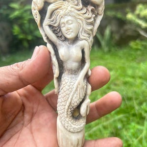 May include: A carved sculpture of a mermaid with flowing hair and a scaled tail. The mermaid is in a relaxed pose, with one arm raised. The sculpture is a light beige color and appears to be made of bone or a similar material.