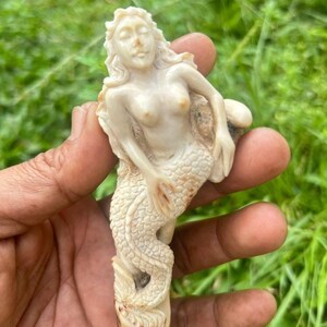 Könnte beinhalten: Eine geschnitzte, elfenbeinfarbene Meerjungfrau-Figur. Die Meerjungfrau hat fließendes Haar, einen ruhigen Ausdruck und eine detaillierte Fischschwanzflosse. Die Skulptur wird vor einem Hintergrund aus grünem Gras gehalten.