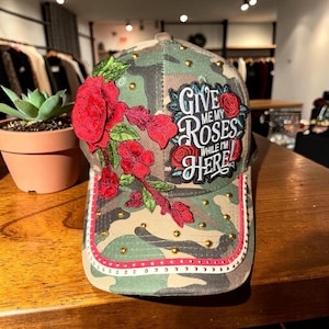 May include: A camouflage baseball cap featuring an embroidered red rose floral design on the left side and a patch on the right side with the text GIVE ME MY ROSES WHILE I'M HERE. The cap is accented with small gold studs and a pink trim.