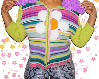 May include: A colorful striped sweater with a large white daisy on the front. The sweater has a pink zipper and a fluffy purple collar.