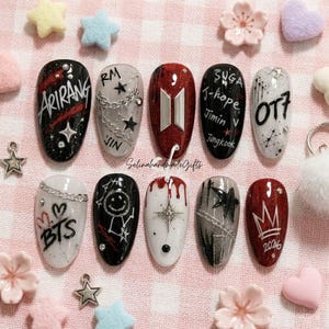 Puede incluir: Un conjunto de diez u&ntilde;as postizas con varios dise&ntilde;os que incluyen el texto Arirang, RM, Jin, Suga, J-hope, Jimin, V, Jungkook, OT7, BTS y 2026. Las u&ntilde;as muestran colores negro, blanco y rojo con detalles met&aacute;licos de cadenas, estrellas y coronas.