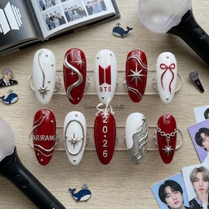 Pode incluir: Um conjunto de dez unhas postiças em vermelho e branco, com detalhes metálicos prateados, berloques de estrela e uma corrente. Os designs incluem o texto BTS, 2026 e ARIRANG, além de um laço vermelho, linhas onduladas e tachas prateadas.