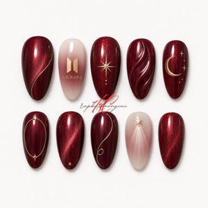 May include: A set of ten almond-shaped artificial nails. The nails feature a deep red color with gold accents, including lines, stars, and a crescent moon. One nail has the text "ARIRANG" and a logo. The nails are arranged on a white surface.