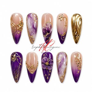 May include: A collection of ten almond-shaped artificial nails with various designs. The nails feature a gradient of colors, including purple, nude, and gold. Some nails have floral and abstract gold accents, while others showcase marble effects.
