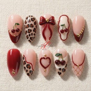 Puede incluir: Una colección de diez uñas artificiales con diversos diseños. Las uñas presentan colores rojo, rosa y marrón, con detalles como cerezas, estampado de leopardo, lazos y corazones. Algunas uñas tienen adornos dorados.