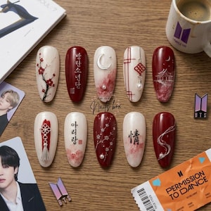 Puede incluir: Un juego de diez uñas postizas con una mezcla de colores crema y rojo intenso con diseños pintados a mano que incluyen flores de cerezo, caligrafía coreana y china, una luna creciente plateada, un nudo tradicional y una pagoda.