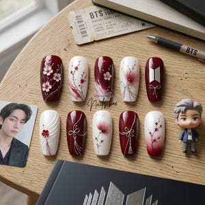 Puede incluir: Un juego de diez uñas postizas con una paleta de colores rojo oscuro y blanco, con diseños florales detallados, detalles de hilos plateados y un logotipo de BTS. El conjunto incluye una tarjeta fotográfica, una entrada y una figura.
