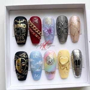 May include: A set of ten artificial nails with various designs. The nails feature gold accents, butterflies, disco balls, and text including "BTS debut", "BE", and "DYNO". Colors include black, red, blue, and pastel gradients.