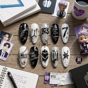 Puede incluir: Un juego de diez uñas postizas con diseños en blanco y negro y detalles plateados, incluyendo los textos Arirang, BTS, 7, We Are Bulletproof y nombres de miembros. Las uñas están decoradas con dijes metálicos, cadenas y formas de estrellas.