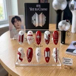 Puede incluir: Un juego de diez uñas postizas exhibidas en un soporte de acrílico transparente. Las uñas presentan diseños en rojo y blanco con detalles plateados, incluyendo el logo de BTS, un micrófono, un corazón, una luna, estrellas y el texto Arirang.