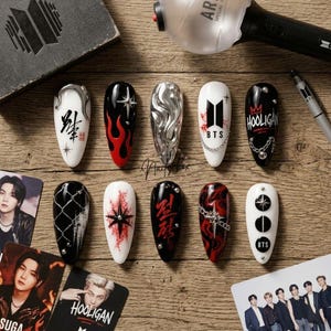May include: A set of ten almond-shaped press-on nails featuring diverse designs including black and red flames, silver metallic textures, chains, star motifs, and the BTS logo. The nails are displayed alongside a light stick and photo cards.