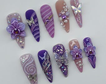 May include: A collection of ten long, almond-shaped artificial nails. The nails feature various designs, including purple and lavender swirls, glitter, butterflies, and floral embellishments. Some nails have pearl and gem accents.