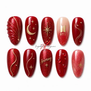 Puede incluir: Un conjunto de diez diseños de uñas rojas. Los diseños incluyen detalles dorados, purpurina y los textos "Ariang" y "BTS". Algunas uñas presentan estrellas, lunas y líneas curvas.