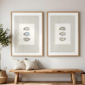 May include: Two framed oyster shell prints. Each print features a light-colored wooden frame, a striped background, and a central rectangular image of three oyster shells. The shells have iridescent colors. The prints hang above a wooden bench with two beige pillows.