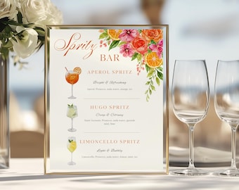 May include: A framed sign with a gold border displays a "Spritz Bar" menu. The menu features watercolor illustrations of Aperol, Hugo, and Limoncello Spritz cocktails, with floral and orange slice accents. Two wine glasses are in the background.