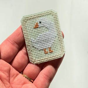 May include: A rectangular cross-stitch patch depicting a white goose with an orange beak and feet. The goose is on a light green background, framed by a light green border. The patch is held in a hand.