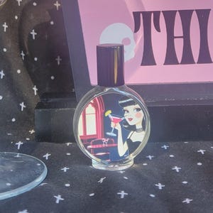 Puede incluir: Una copa de martini llena de una bebida de color marrón claro se encuentra junto a un letrero rosa que dice "YOU Goth THIS". También hay un pequeño frasco de perfume con una mujer sosteniendo un cóctel. El fondo es una tela negra con cruces blancas.