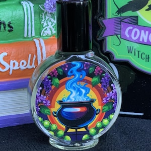 May include: A clear, round glass bottle with a black cap, featuring a colorful label with a cauldron and blue flames. The label is surrounded by purple grapes and green orbs. The bottle is themed for Halloween or a witchy aesthetic.