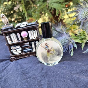 May include: A clear, round perfume bottle with a black cap, featuring a vintage illustration of a ballerina. A miniature bookshelf with an owl figurine sits nearby. The background includes blue thistle flowers and greenery.
