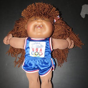 May include: A doll with brown yarn hair and a tan complexion wearing a blue athletic outfit. The top features the text "OlympiKids 1996 Official Team Mascot USA" with the Olympic rings logo. The outfit includes blue shorts with red and white trim.