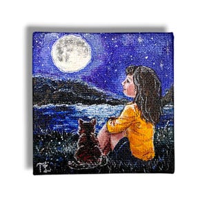 May include: A small square painting depicts a girl and a cat gazing at a full moon in a starry night sky. The girl wears a yellow shirt and sits with the cat in a grassy area near water. The painting has a whimsical, dreamlike quality.