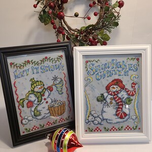 May include: Two framed cross-stitch pictures with snowmen, one in a black frame and the other in a white frame. The black framed picture says "Let it Snow!" and the white framed picture says "Snowflakes Glisten". A red and silver Christmas ornament is in front of the pictures.
