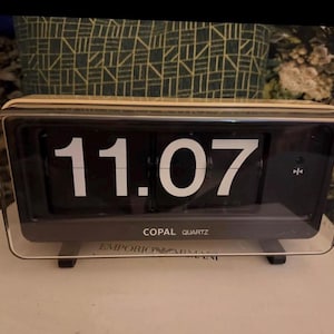 May include: A vintage Copal Quartz flip clock displaying 11:07. The clock has a black face with white numerals and a clear plastic casing. The words "Copal Quartz" are printed on the face. The clock is on a white surface.
