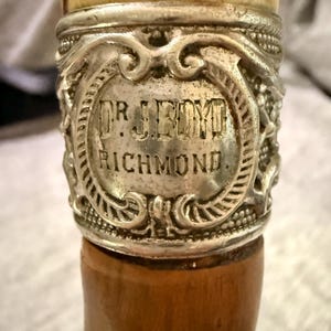 Puede incluir: Primer plano de un mango de bastón. El mango presenta una banda plateada con el texto grabado "DR. J. BOYD RICHMOND" dentro de un marco ornamentado. El mango es de un material marrón claro, y la parte superior es de un color más claro.