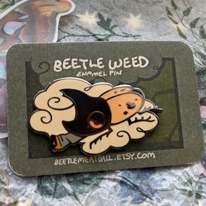 May include: An enamel pin featuring a stylized character with a black hood and orange face, surrounded by cream-colored cloud shapes. The pin is attached to a rectangular olive green backing card with the text BEETLE WEED ENAMEL PIN.
