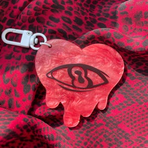 May include: A red heart-shaped keychain with a black eye design. The keychain has a white clasp and a dripping effect at the bottom. The background is a red fabric with a black leopard print.