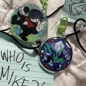 May include: Two round acrylic keychains with cartoon character designs. One features a black figure with a red bow tie, the other a green and blue character. Both have green clasps and black cords. A blue paper with the text "WHO IS MIKE?!" is also visible.