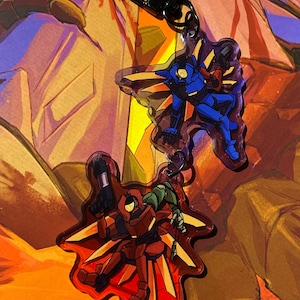 May include: Two acrylic keychains featuring stylized robotic figures. One figure is blue with yellow wing-like accents, and the other is red with yellow wing-like accents. Both figures hold small black weapons and are attached to a black metal clasp.