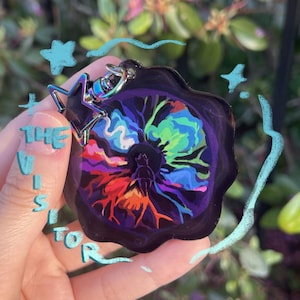Puede incluir: Un llavero acrílico colorido con un cierre en forma de estrella. El llavero presenta un diseño vibrante con el texto "THE VISITOR" y una figura central rodeada de colores arremolinados de azul, verde, naranja y rojo.