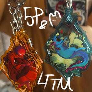 Op de afbeelding: Twee acryl sleutelhangers met zilveren kettingen. Eén is oranje met een rode figuur die een instrument bespeelt. De andere is blauwgroen met een blauw wezen. De sleutelhangers hebben de tekst "5P&" en "LTTM".