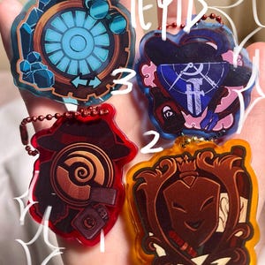 May include: Four acrylic keyrings with various designs. One features a blue and brown gear, another has a blue and white emblem, a third is a red and brown design, and the last is a brown and yellow emblem.