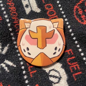 Pode incluir: Um pin redondo, laranja e branco, com um design semelhante a um gato. O pin apresenta uma cruz dourada no centro, marcas rosadas nas bochechas e pequenos pontos pretos. O pin está sobre um fundo com padrão em preto e branco.