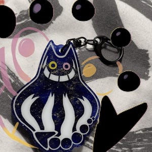 Puede incluir: Un llavero azul con forma de gato con una cara sonriente, ojos amarillos y rosas y garras blancas. El llavero tiene un cierre y una cadena negros. El cuerpo del gato está lleno de purpurina dorada.