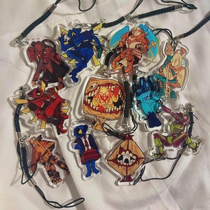 May include: A collection of clear acrylic charms featuring various cartoon characters in vibrant colors. Each charm has a black lanyard attached with a silver clasp. The characters include robots, monsters, and figures in action poses.