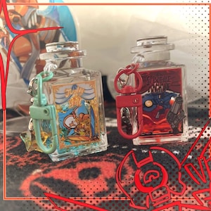 May include: Two clear rectangular bottle-shaped keychains with metal clasps. The left keychain has a light green clasp and a label reading "Judge of Hell" with a character illustration. The right keychain has a red clasp and a label reading "Supreme Machine" with a robotic character illustration.