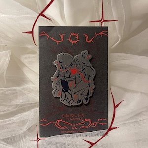 May include: An enamel pin featuring two stylized figures holding a red heart, set against a dark gray background. The pin is on a dark gray card with red decorative elements and text that reads "ENAMEL PIN BY MIPEXCH" and a website address.