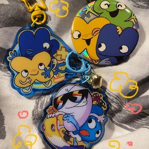 Peut inclure: Une collection d'accessoires colorés sur le thème des personnages. Comprend un porte-clés en forme d'étoile jaune, deux porte-clés de personnages et un badge rond. Les porte-clés présentent des personnages bleus et jaunes avec des dessins de style dessin animé.