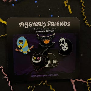 以下が含まれることがあります： Mystery Friendsと書かれた黒い台紙にセットされた4つのエナメルピン。青と黄色の生き物、オレンジ色の目を持つ黒い幽霊のような姿、白い頭蓋骨のような顔、そして黒い猫など、デフォルメされたキャラクターが描かれたピンバッジのセット。