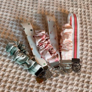 May include: Four fabric pacifier clips in various patterns. One clip is green plaid, another has polka dots, and two feature floral designs. Each clip has a metal clasp and a small metal charm.