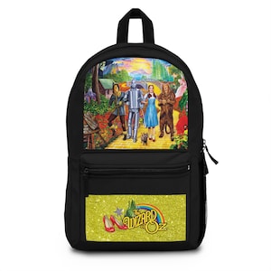 May include: A black backpack featuring a colorful graphic of The Wizard of Oz characters on the upper panel and a yellow glittery panel below with red slippers, a wand, a rainbow, and the text The Wizard of Oz printed in a stylized font.