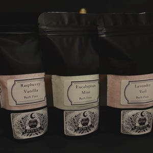 Peut inclure: Trois sachets noirs de sels de bain avec des étiquettes. Les étiquettes indiquent "Raspberry Vanilla", "Eucalyptus Mint" et "Lavender Veil". Chaque étiquette porte la mention "Bath Fizz" et le logo Ritual Bath Co. Les sachets sont sur une surface sombre.