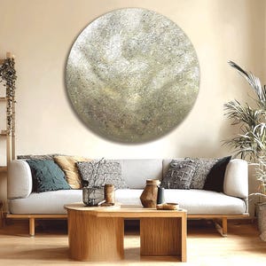 Puede incluir: Una gran obra de arte abstracta redonda con una superficie texturizada y moteada en tonos grises, beige y dorado cuelga sobre un sofá gris claro. El salón incluye una mesa de centro de madera, cojines decorativos y una planta.