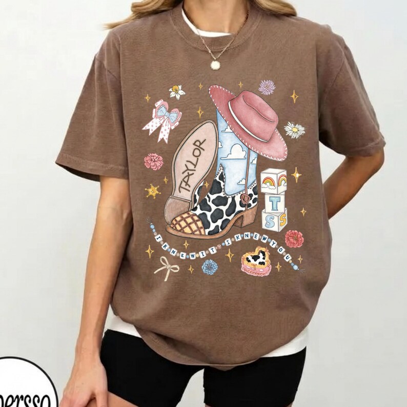 Op de afbeelding: Een bruin t-shirt met korte mouwen en een grafische print van een cowboylaars met koeienprint, een roze hoed, alfabetblokken met de letters TS en de tekst TAYLOR. Het ontwerp bevat kleine sterretjes, bloemen en de tekst I KNEW YOU.