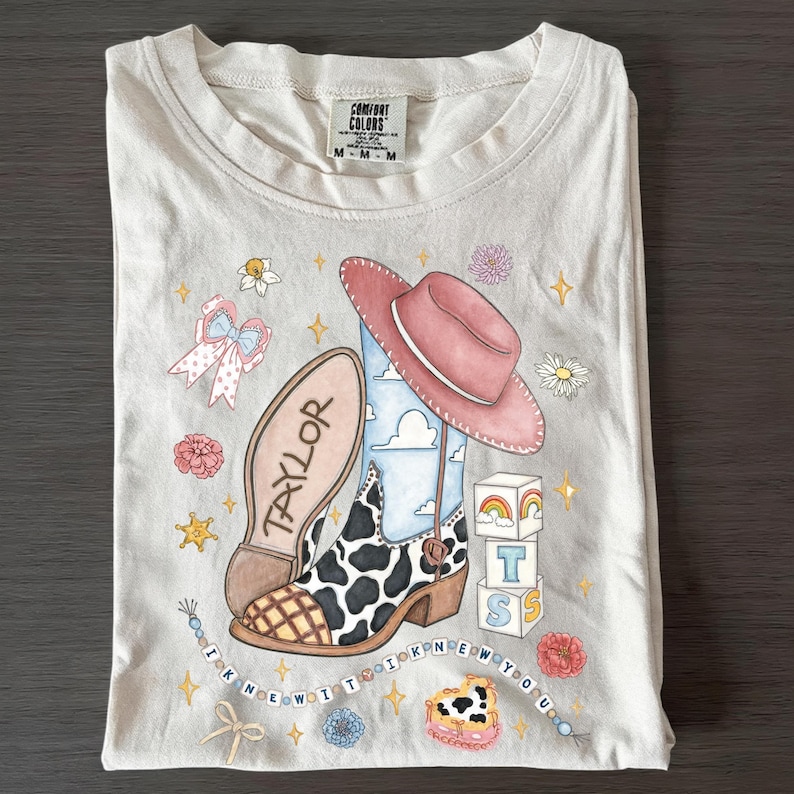 Op de afbeelding: Een beige t-shirt met een grafische print van een cowboylaars met koeienprint, een roze hoed, een strik, bloemen en blokken met de letters T S. De tekst TAYLOR staat op de zool en een kralensnoer bevat de tekst I KNEW YOU WERE TROUBLE.