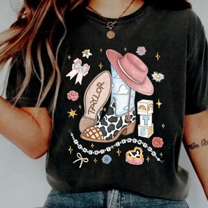 Op de afbeelding: Een antracietgrijs t-shirt met een grafische print van een cowboylaars met koeienvlekken, een roze hoed, een strik en blokken met de letters T, S en een regenboog. De zool is gelabeld met TAYLOR en de tekst I KNEW YOU staat in een boog.